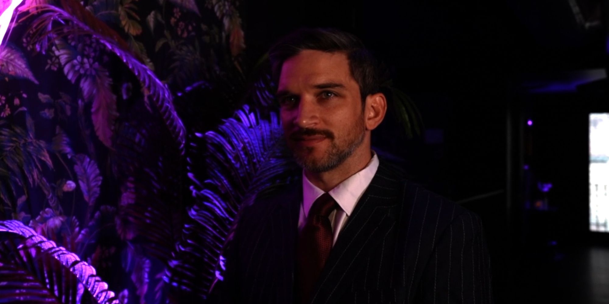 O ator Evan Jonigkeit como Raphael, vestindo um terno e gravata e de pé contra um cenário floral em atrabilioso.