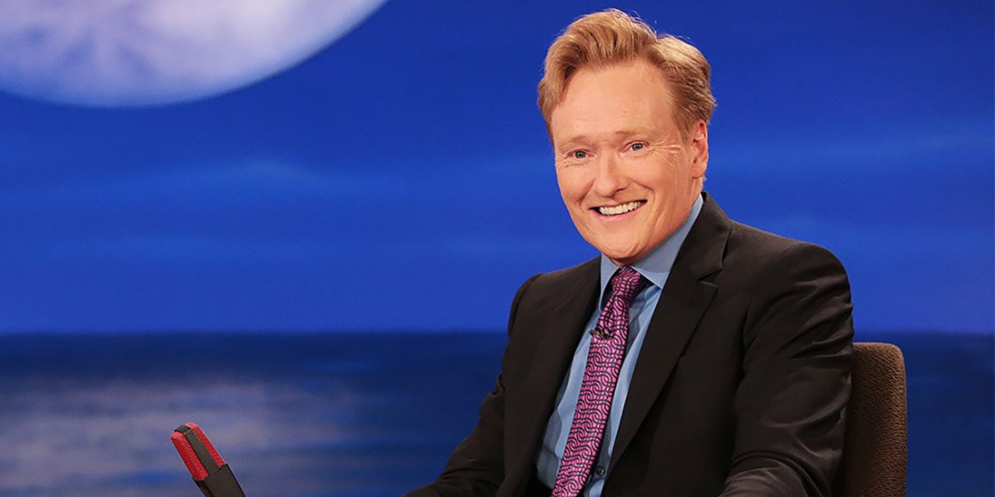 Conan O'Brien sorrindo atrás de sua mesa na noite com Conan O'Brien.