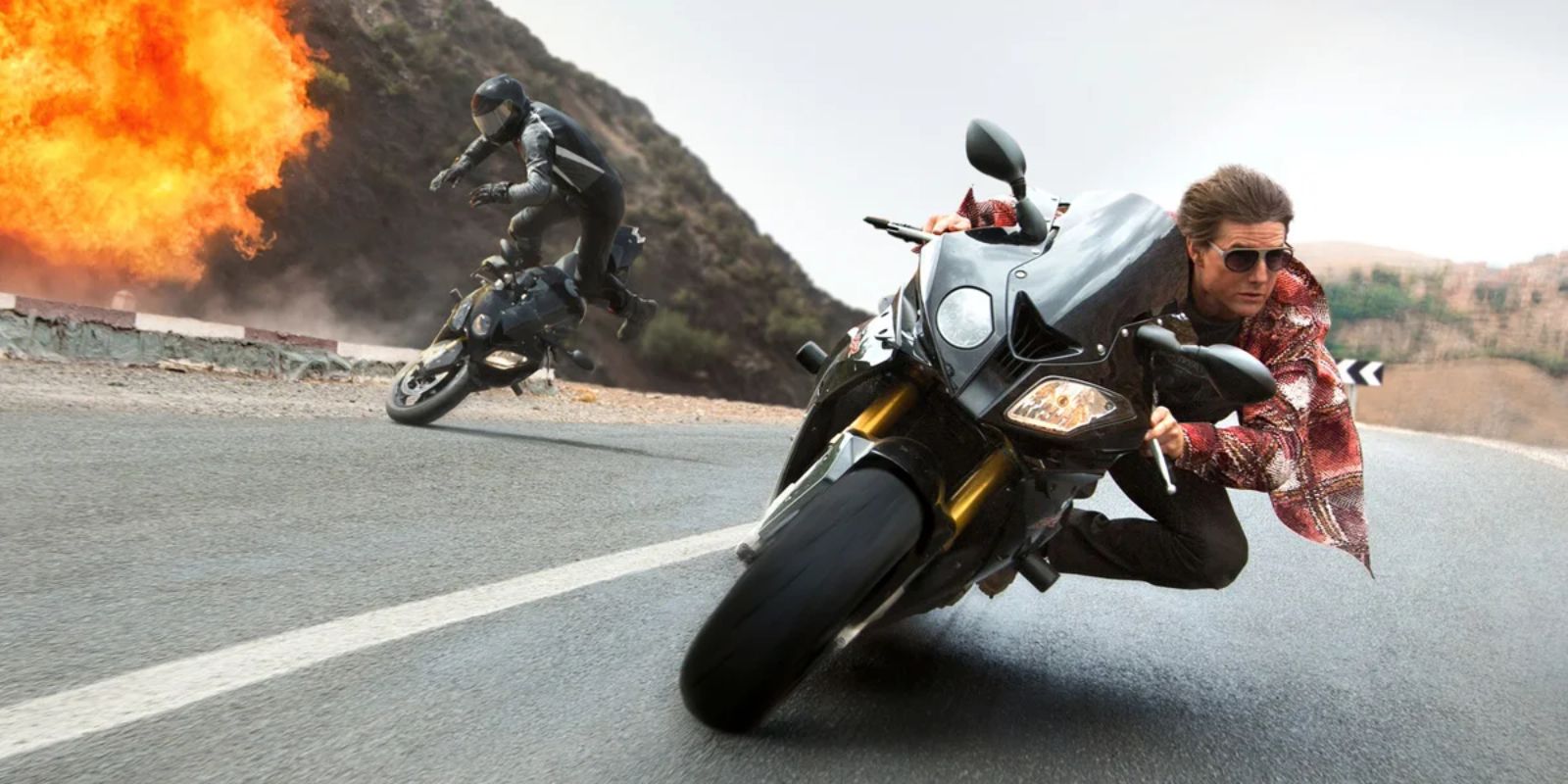 Ethan Hunt (Tom Cruise) riding a motorcycle with an explosion and crash in the background in Mission: Impossible – Rogue Nation (2015)