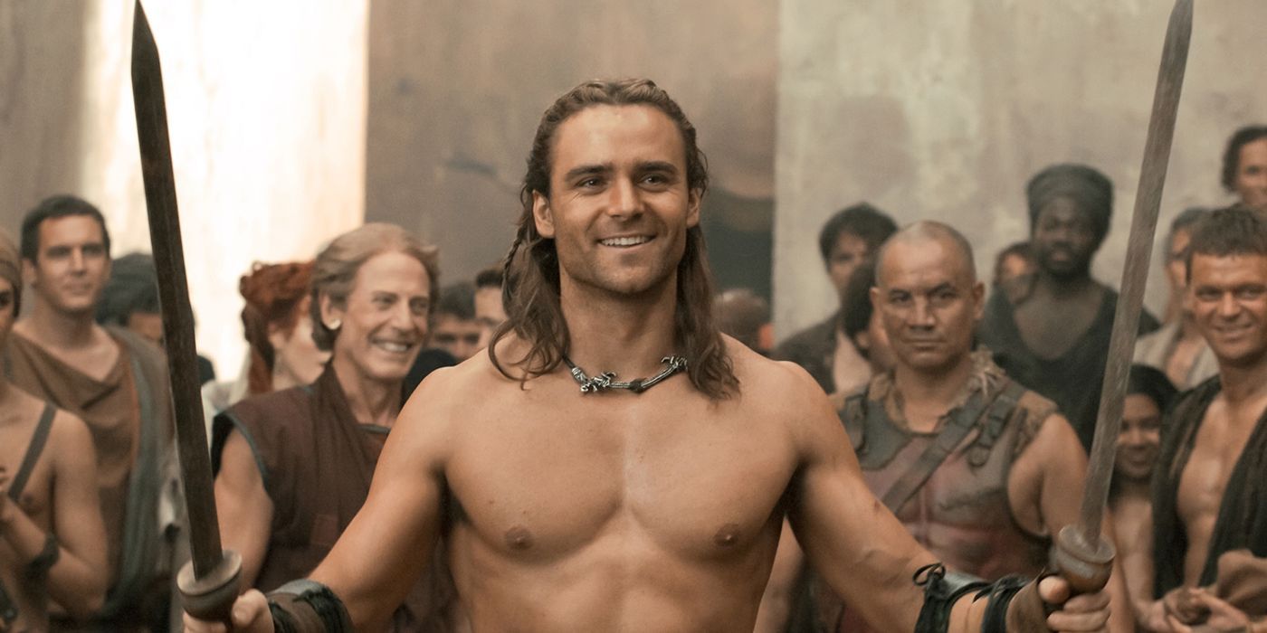 Dustin Clare as Gannicus smiling and holding swords with a crowd behind him in Spartacus: Gods of the Arena.