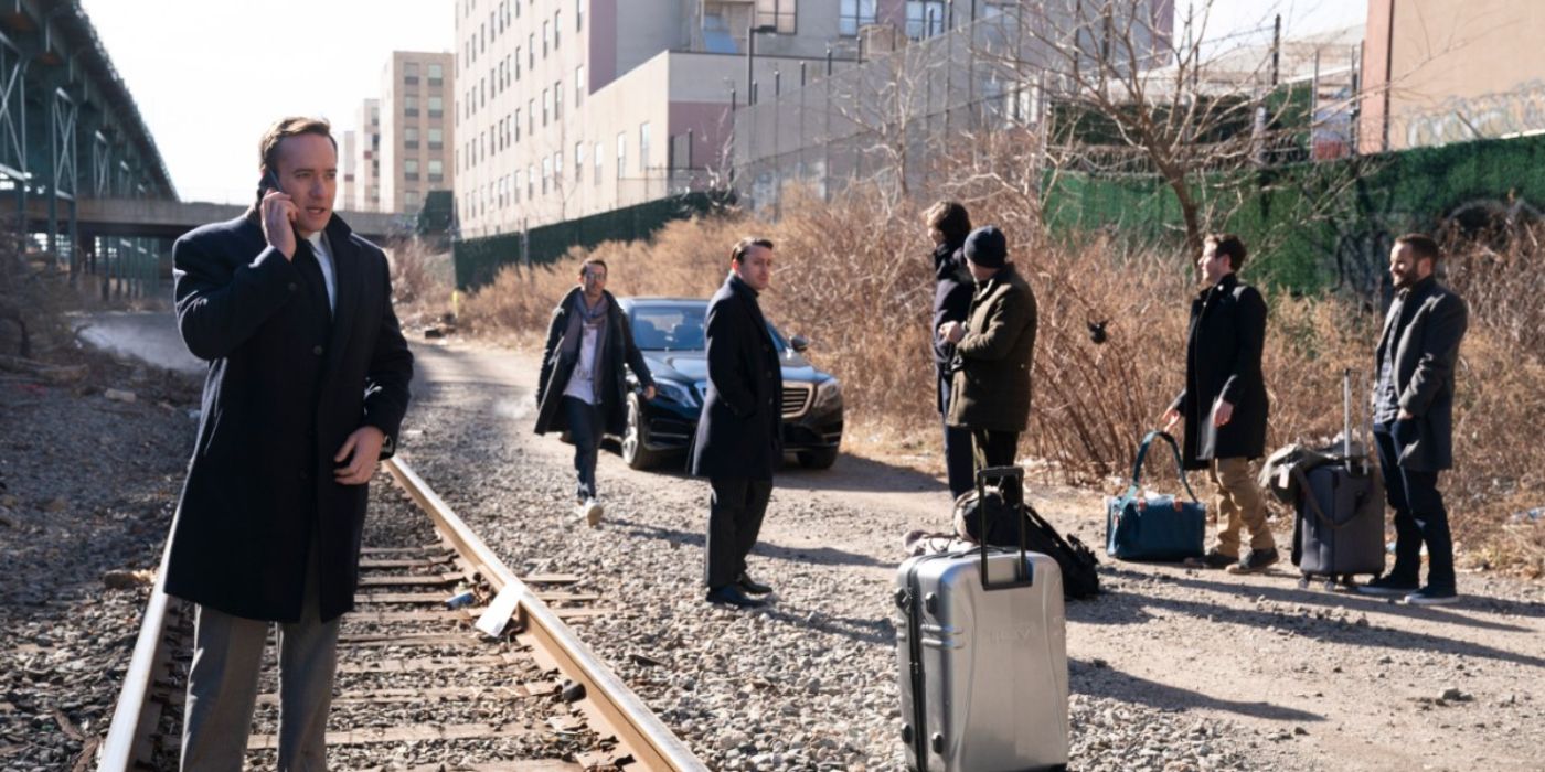 Tom on the phone while standing on train tracks while Roman, Kendall, Connor, and their entourage stand behind him