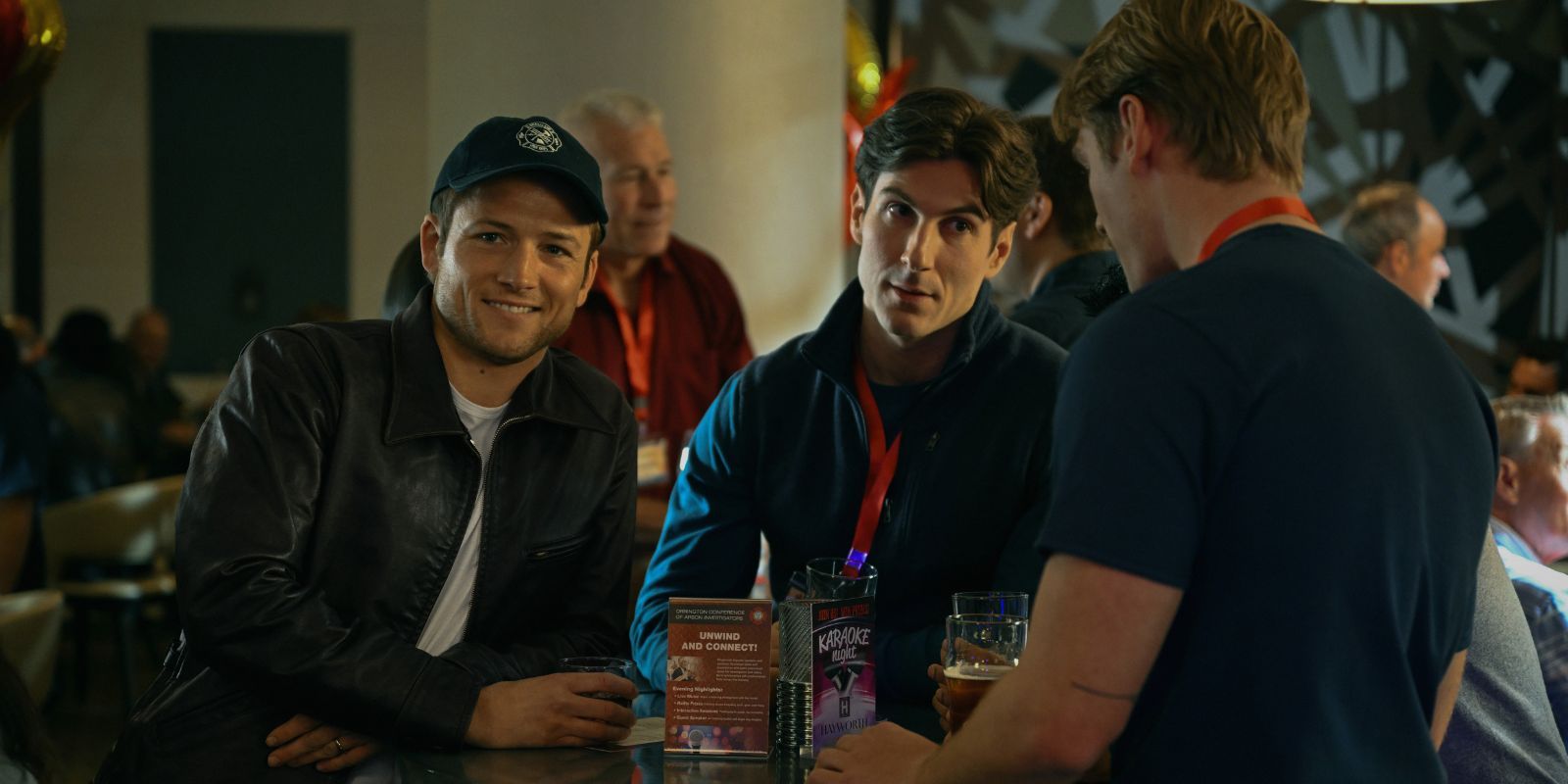 Taron Egerton sorrindo enquanto dois jovens conversam em fumaça