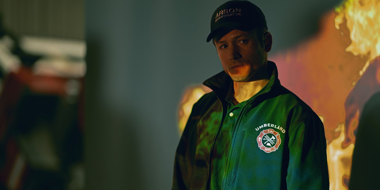 Taron Egerton em pé na frente de uma tela com fogo na fumaça
