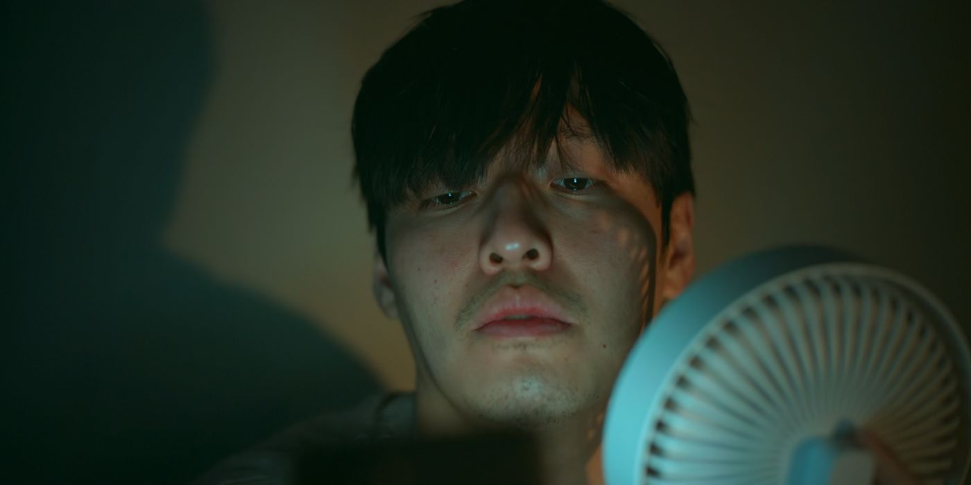 Woo-sung's face is lit by his phone's light while he checks the crypto market, as he points a fan towards his face in the dark to save money in Netflix's Wall to Wall