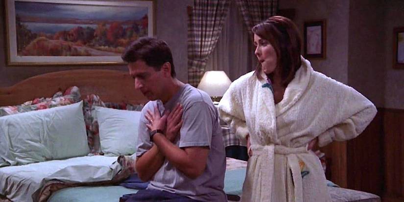 Ray sits on a bed with hands over his chest as Debra stands behind, hands on hips in Everybody Loves Raymond.