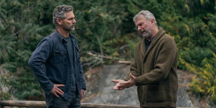 Eric Bana and Sam Neill as Kyle and Paul wearing a blue shirt and green sweater standing in front of a waterfall in 'Untamed.'