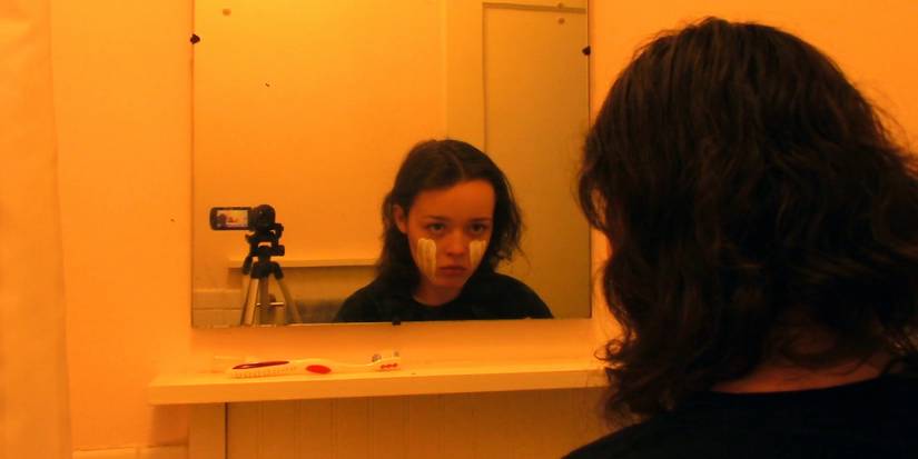 Jauna sieviete, kas skatās uz viņas pārdomām spogulī ar videokameru blakus viņai, mēs visi ejam uz pasaules gadatirgu (2021)