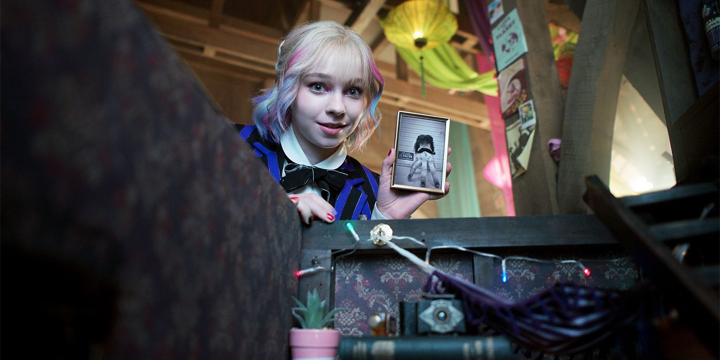 Emma Myers as Enid holding up a vintage photo of Thing and looking in a chest in Wednesday Season 2.