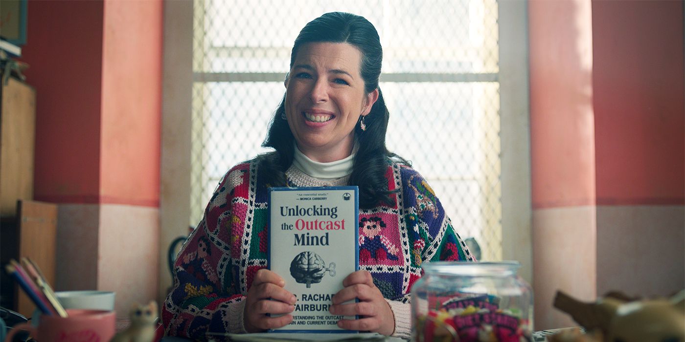 Heather Matarazzo holding a book titled "Unlocking the Outcast Mind" and smiling in Wednesday Season 2.