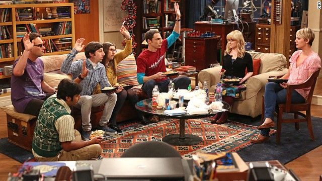 The Big Bang Theory cast sitting in the living room raising their hands